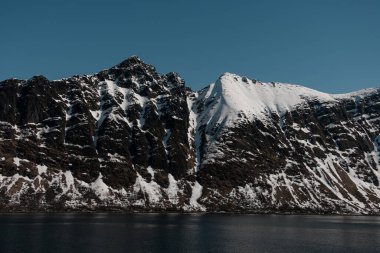 Tepesi karla kaplı yüksek kayalık dağlar. Mavi deniz dağın eteğinde, parlak mavi bir gökyüzünün arka planında yer almaktadır.
