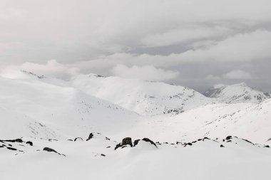 Güzel karla kaplı dağ kitlesi, beyaz bulutların üzerinde süzüldüğü ve karla kaplı dağların tepelerine tutunduğu gökyüzünün arka planına karşı.