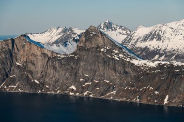 Dağın kayalık yamacının eteğindeki mavi deniz ardında pek çok keskin tepenin görülebildiği mavi gökyüzü vardır.