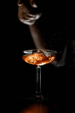 Üzerinde bir adam eli olan, karanlıkta bardakta sallanan bir kokteyl olan saplı bir bardağa odaklan.