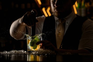 Barmen kokteyli limon dilimleri ve nane yapraklarıyla dikkatlice karıştırıyor. Böylece damlalar farklı yönlere uçuyor.