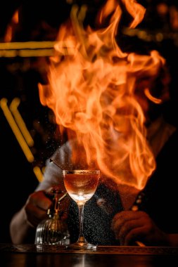 Barın tezgahında kahverengi bir karışım ve zeytin şişiyle dolu yüksek saplı bardaklar var. Barmen bu bardağa ateş püskürterek havaya yanıcı sıvı püskürtüyor.