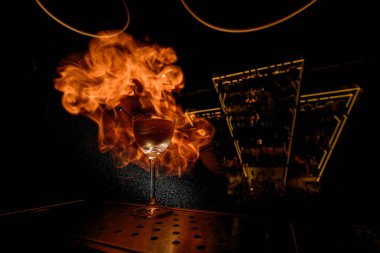 Glass filled with a cocktail stands on the bar counter against the background of drops of spray that catch fire so that even the bartender cannot be seen behind the flames against the background of an clipart