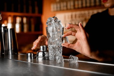 Kaydıraklı cam karıştırıcı buz küpleriyle doludur. Böylece barın üzerine dökülür ve barmen kadınlar tarafından alınıp bulanık bir arka planda eritilirler.