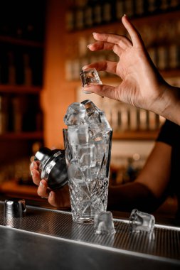 Barmenin kadın eli bir buz küpü gösteriyor, ve diğer elinde de buz küpleriyle dolu bir kaydırağın çelik kapağını tutuyor, böylece barın tezgahına dökülüyorlar.