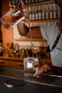 Kadın barmen elinde buzlu kokteyl bardağı, içine kahverengi kokteyl dökerek bardağı barın üstüne kaldırıyor.