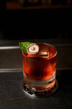 Yuvarlak bir metal standın üzerinde yeşil bir yaprak ve mantar kapağıyla süslenmiş buzla birlikte kahverengi bir kokteyl var.