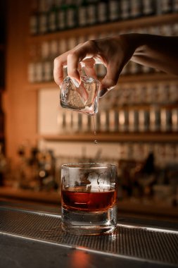 Kahverengi içki bardağın içinde, yarısı dolu bir kadın eli üzerinde büyük bir buz küpü tutuyor ve damlalar suya dökülüyor.