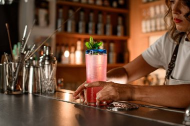 Kadın barmen, bir bar vitrininin bulanık arka planında, üzerinde nane yaprakları ile süslenmiş pembe bir kokteyl ile uzun bir bardak tamir eder.