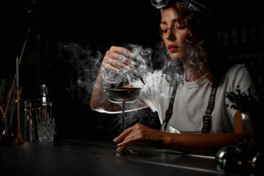 Kadın barmen, yanan odun parçacıklarıyla etrafa duman üflüyor.