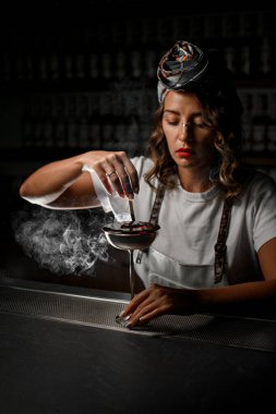 Elinde odun parçasıyla barmen bir kadın için için için için için yanan ve arka planda bulanık duran metal saplı bir camın etrafında duman üfleyen.