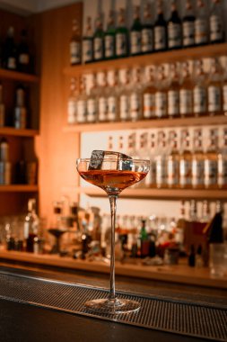 Bir bardağın uzun, ince gövdesine yakın plan, yarısı büyük bir buz küpüyle dolu bir kokteyl, tezgahın üzerinde, aydınlatılmış bir bar vitrininin arka planında çeşitli alkollü içeceklerle birlikte
