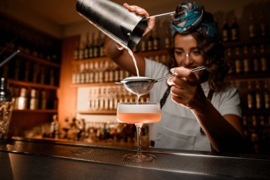 Sevimli bayan barmen, süngerin içine köpüklü bir içecek doldurur. Bir kokteyl, bir bar tezgahının üzerinde duran saplı bir bardağa sıkıştırılır.