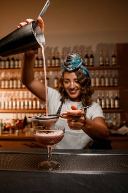 Mutlu gülümseyen bayan barmen, süzgecin üzerinden ince saplı bardağa tutarak sallanan köpüklü kokteyli doldurur.