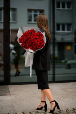 Yansıtıcı binanın yanında, esmer bir kadın camlara bakar, büyük bir buket kırmızı gül tutar, kentsel aynalı güzellikten büyülenir.