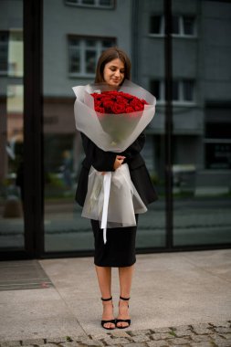 Neşeli gülüşü ayna binasına yansıyor, kocaman bir buket kırmızı gülü kucaklıyor..