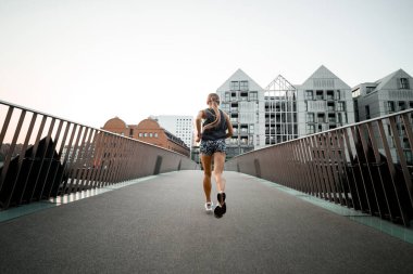 Kısa şortlu ve kolsuz tişörtlü genç bir kadının kaldırımda şehrin arka planındaki yüksek binalara doğru koşuşunun arka görüntüsü.