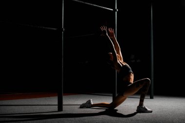 Spor salonunda tek dizinin üzerinde duran kaslı kadın, iki elini kaldırıyor, kolunu kaldırıyor ve sırtını eğiyor.