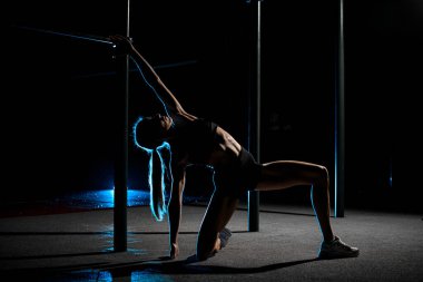 Genç zayıf kadın spor salonunda esneme egzersizleri yapıyor, bir bacağı dizinde, diğer bacağı yanda duruyor, ve bir kolu başının üstüne doğru uzanıyor.