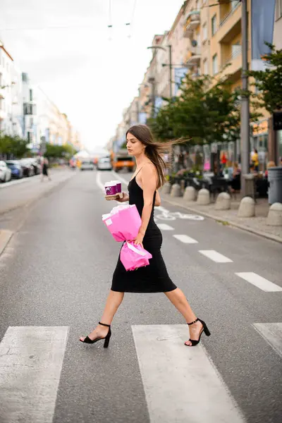 Elinde güzel bir buket çiçek ve iki bardak sıcak içecekle caddeden karşıya geçen narin bir kadın.