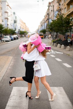 Siyah beyaz elbiseli iki genç kız birbirlerine sarılıyorlar. Bir şehir caddesinde ellerinde çiçekler ve bir bardak kahveyle.