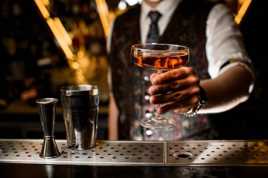 Erkek barmen, elinde tutarken kahverengi bir kokteyl ile dolu saplı bir şarap bardağını gösteriyor ve bir barın üstündeki metal bardak ve jigger ile kameraya uzatıyor.