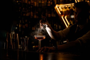Barmen bardağa kahverengi kokteyl püskürtür, bar tezgahında bulanık dekoratif lambalar ve bar vitrinleriyle