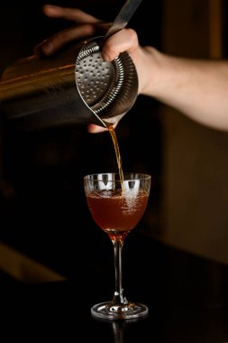 Erkek bir barmenin eli, şarap bardağına kahverengi bir içecek dökerken, baş parmağıyla tuttuğu buz süzgecinin içinden süzülüyor.
