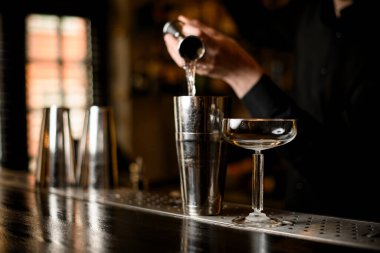 Erkek barmen elleri metal kokteylden buhar kokteyline temiz sıvı döker, uzun saplı kokteyl bardakları yakınlarda durur.