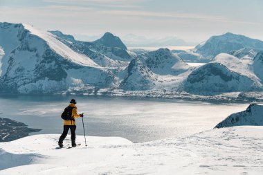Tek elli bir kayakçı dağın tepesine ulaşmış, uçsuz bucaksız dağ ve deniz manzarasına hayran kalmış, keskin bir uçurumun kenarında duruyor.