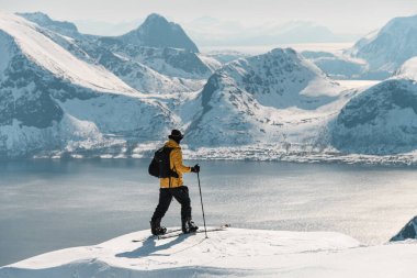 Fotoğraf, tek kollu mutlu bir kayakçının arka planında, tepenin hemen kenarında, bir dağın tepesinde duran deniz manzarası ve önü açık dağların karşısında duran bir kayakçının fotoğrafı.