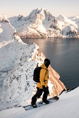 Genç tek kollu kayakçı neredeyse tepeye yakın karlı bir yamaçta duruyor ve merkezinde bir parça deniz olan güzel bir dağ yayı manzarasına bakıyor.