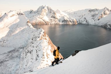 High angle view of a beautiful mountain range with a small piece of sea in the center, a splitboarder standing on his splitboard in the foreground and admiring the beauty clipart