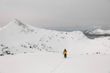 Tek kollu kayakçı dağların arasında asfalt bir kayak üzerinde yürüyor, arkasında keskin bir tepe ve küçük bir nehir ve dağlarla birlikte yüksek bir dağ bırakıyor.