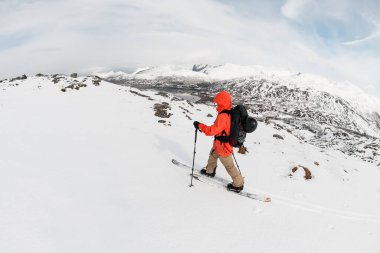 Turuncu ceketli bir kayakçının üst görüntüsü karla kaplı bir dağ sırasının arka planında aralarından akan küçük bir nehir ile kayak yapıyor.