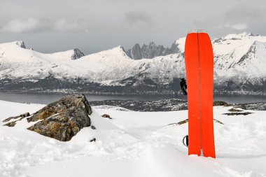 Orange splitboard stands in the snow on the background of a sea bay formed between the mountains. Winter landscape in the mountains clipart