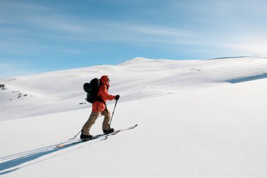 Turuncu ceketli kayakçının yan görüntüsü kayakların üzerinde başka bir tepe ve mavi gökyüzünün arka planına doğru yumuşak, karlı bir dağ yamacına doğru ilerliyor.