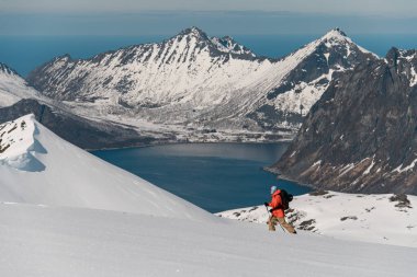 Mavi arka planda yüksek açılı güzel bir sıradağ manzarası. Ön planda, bir kayakçı karlı bir yamaçta kayak yapıyor, kayağın eteğinde bir deniz körfezi var.