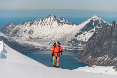 Yüksek bir dağdaki kayak tırmanışını tamamlayan bir kayakçının, yüksek dağların ve denizin bulanık arka planına yakın görüntüsü.