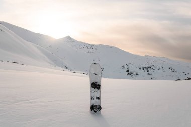 White splitboard stands in the snow against the background of a snowy plain and mountains with sharp peaks, behind which a bright spring sun peeks out in a light sky with white clouds clipart