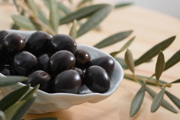 Detail of a ceramic bowl with black olives next to some olive branches on a wooden table