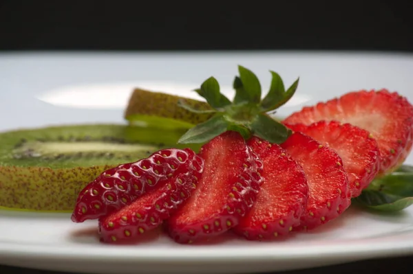 Image of a white plate with a sliced strawberry next to two slices of kiwi with skin next to fresh strawberry leaves