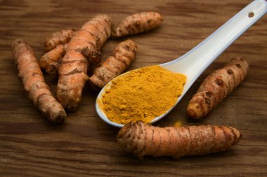 A spoon of turmeric (Curcuma longa) powder surrounded by rhizomes of the same plant on a wooden board