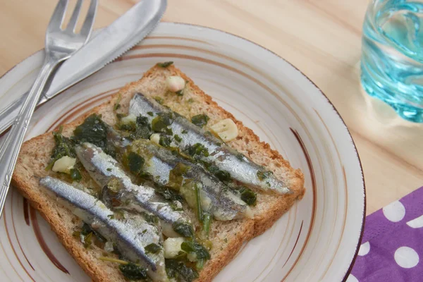A slice of bread with anchovies seasoned with oil, vinegar, garlic and parsley on a plate with cutlery seen from above