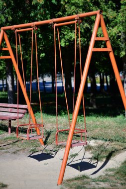 Güneşli bir günde ağaçlarla çevrili sessiz bir parkta salıncaklar çocukları oyun oynamaya ve dışarıda eğlenmeye davet ediyor.