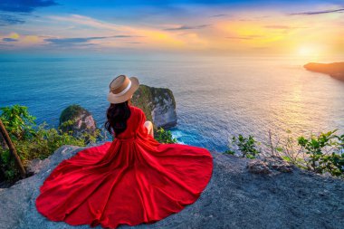 Uçurumda oturan ve Endonezya 'nın Nusa Penida adasındaki Kelingking Sahili' nde gün batımını izleyen bir turist..