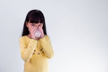 Küçük sevimli Taylandlı oyuncu kız beyaz arka planda izole edilmiş bir bardak süt tutuyor ya da içiyor.