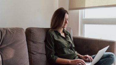Young blonde woman using laptop at home. Beautiful caucasian girl smiling, sitting on sofa and working remotely with notebook