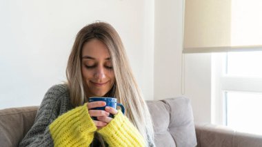 Woman wearing sweater warming her hands with hot drink. Portrait of blonde woman at home