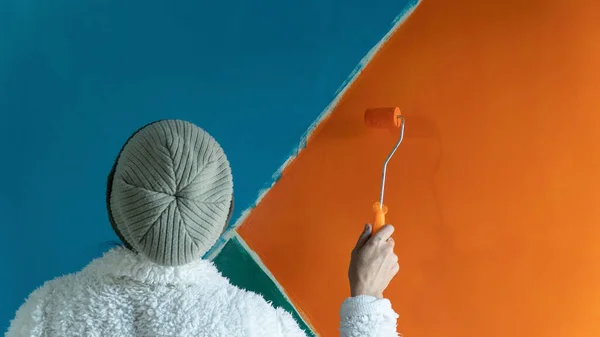 Middle aged woman painting a wall. Woman in winter clothes painting wall with roller brush. Moving house. Selective focus included. Noise and grain included